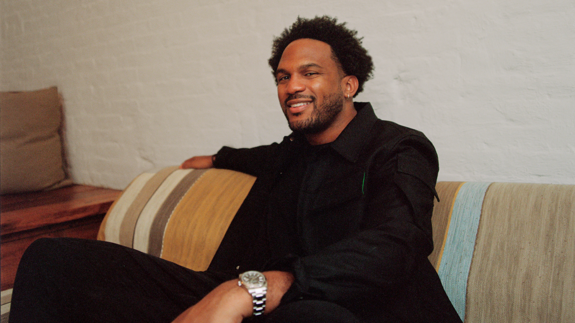 Kickstarter CEO Everette Taylor, wearing all black, sits on a couch and rests his left arm on his left knee. A watch is visible on his wrist.